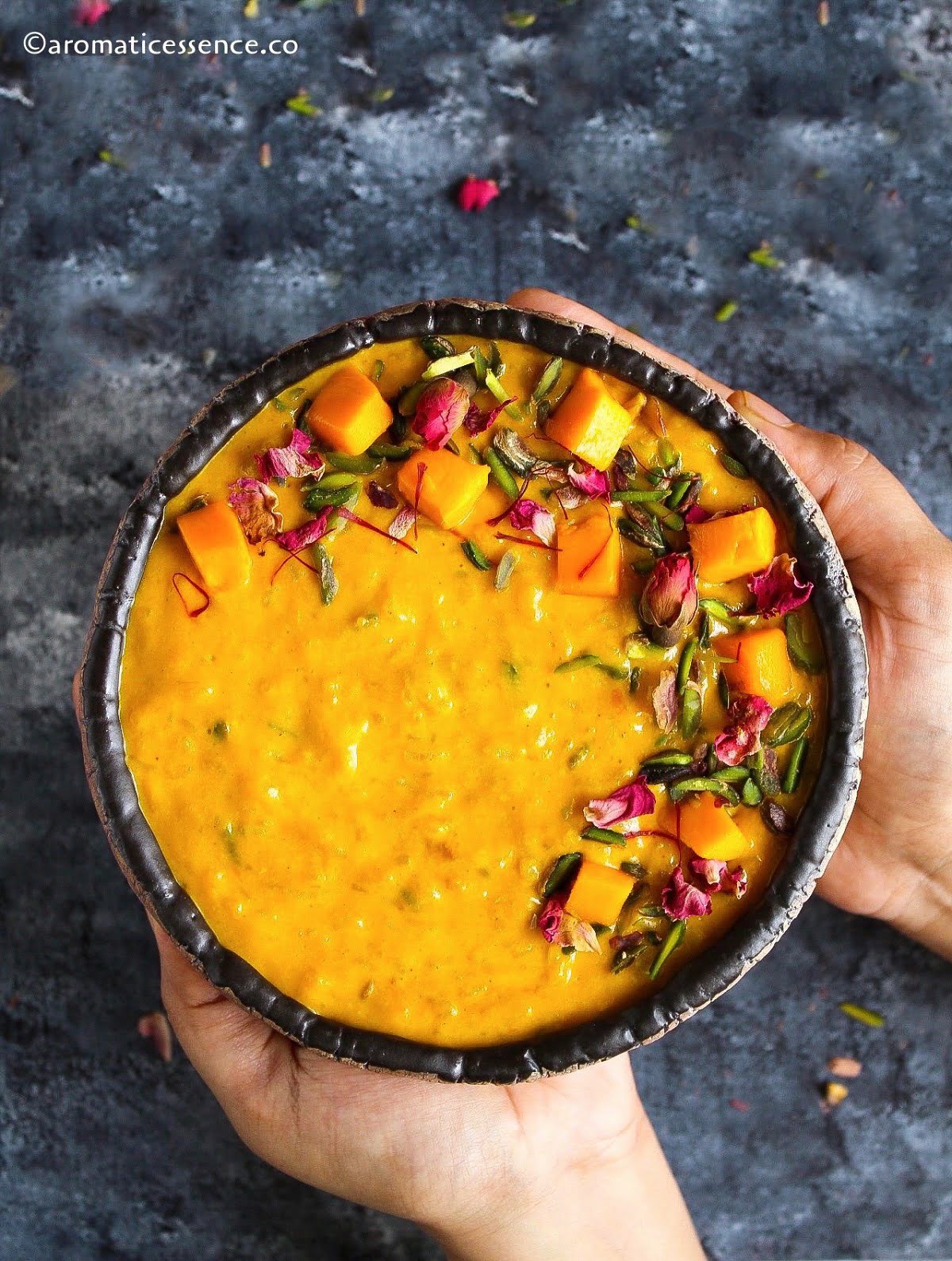 Hands holding a bowl of mango rice kheer against a dark background