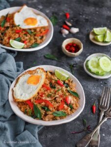Spicy basil fried rice served in two shallow bowls alongside lime wedges, Prik nam pla, and sliced cucumber