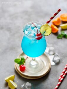 45 degree angle shot the blue lagoon mocktail garnished with lemon wheel, maraschino cherry, and cocktail umbrella