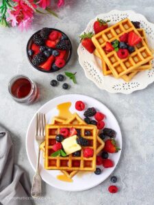 Eggless waffles topped with butter and berries, drizzled with maple syrup served on white plates