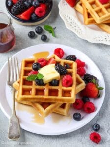 45 degree angle shot of egg-free waffles topped with butter, berries, and maple syrup on a white plate
