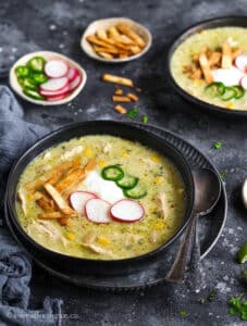 Green chile enchilada chicken soup served in two black bowls, topped with radish slices, jalapenos, sour cream, and tortilla strips