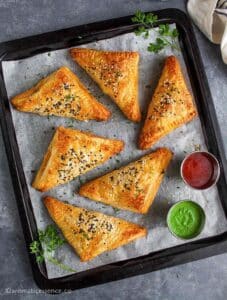 Six paneer puffs on a black baking tray served with ketchup and cilantro-mint chutney