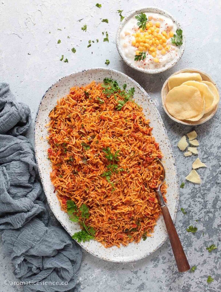 tomato rice served in a white speckled platter with boondi raita and papadum