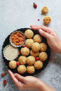 Hands holding a round metal tray containing til ke laddu